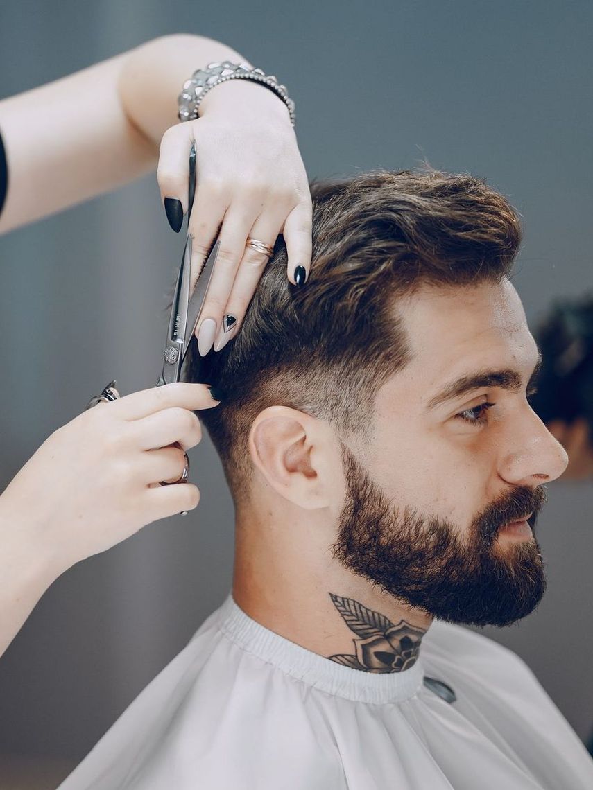 Barber cutting a man's hair, using scissors. Man has dark beard and neck tattoo, wearing a cape.