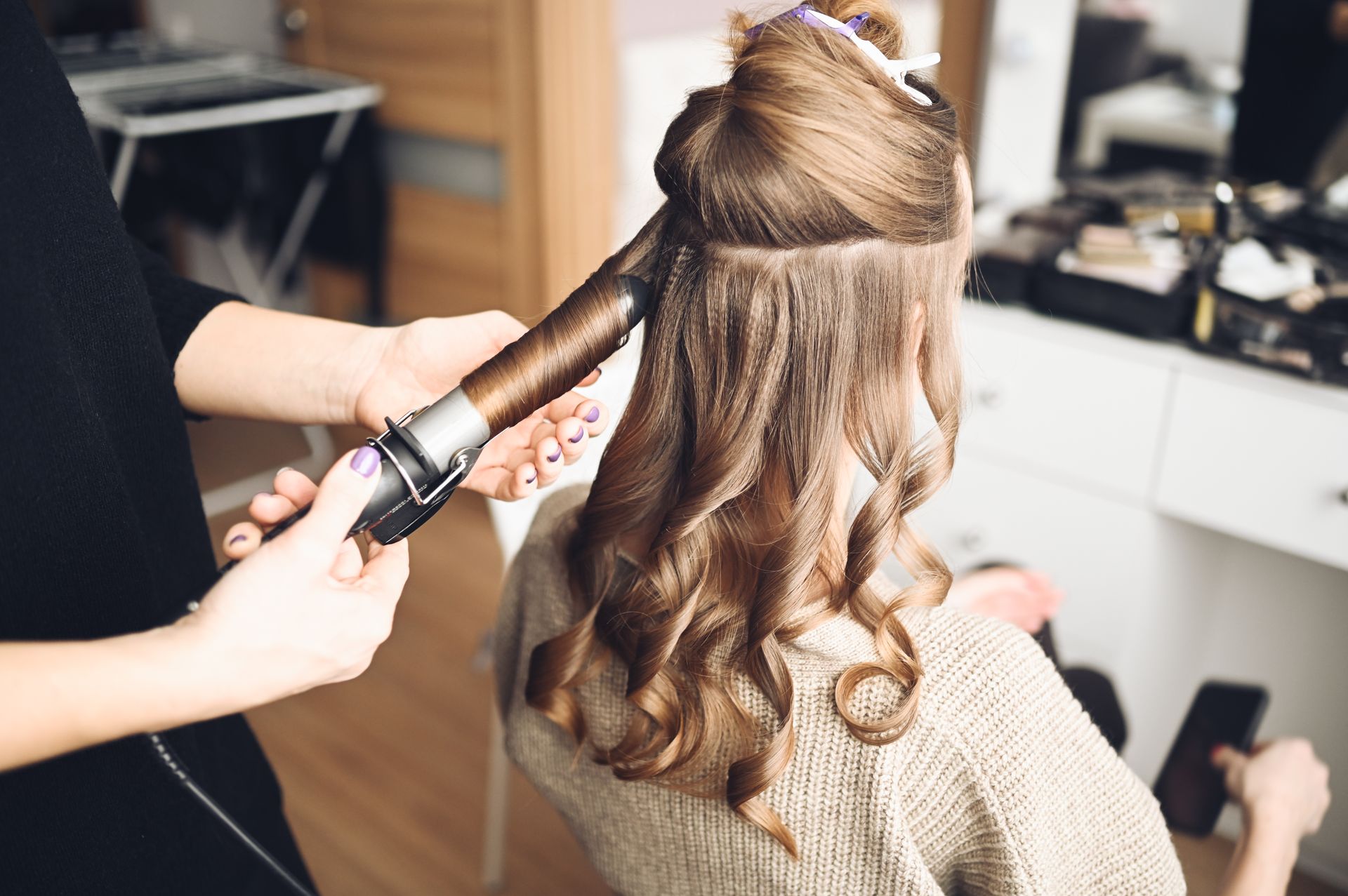 A hair stylist uses a curling iron to style long brown hair in a salon setting.