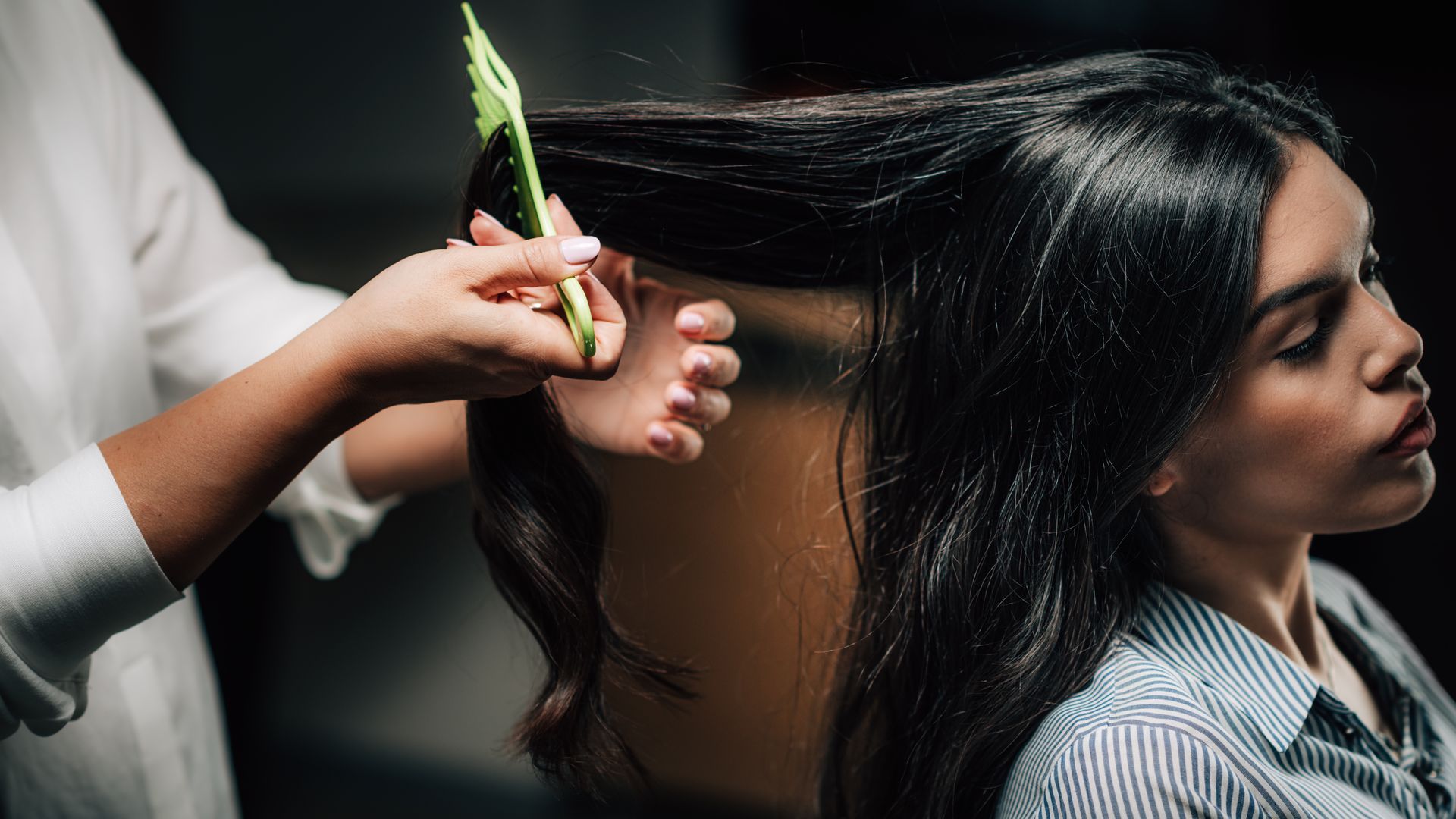 Hairdresser combing a client's long, dark hair. The client sits while the hairdresser holds a green comb.