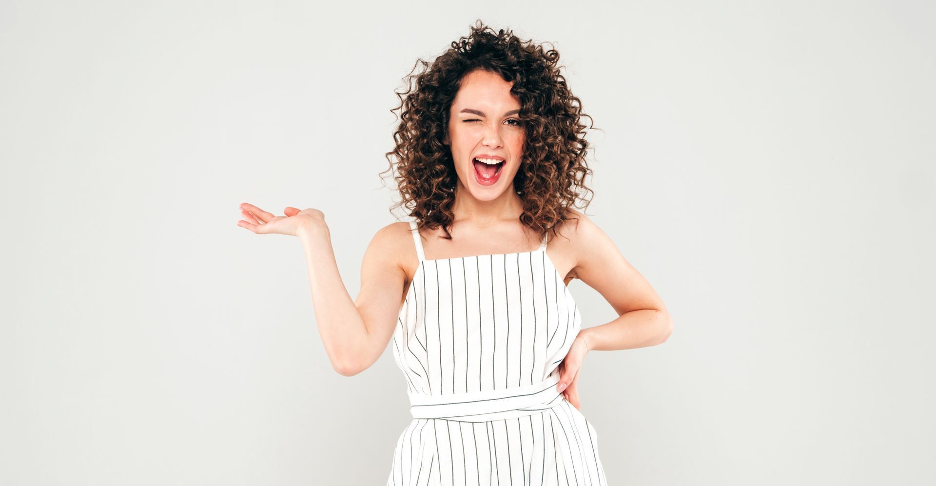 Woman with curly hair in a striped dress winks and gestures with her hand.