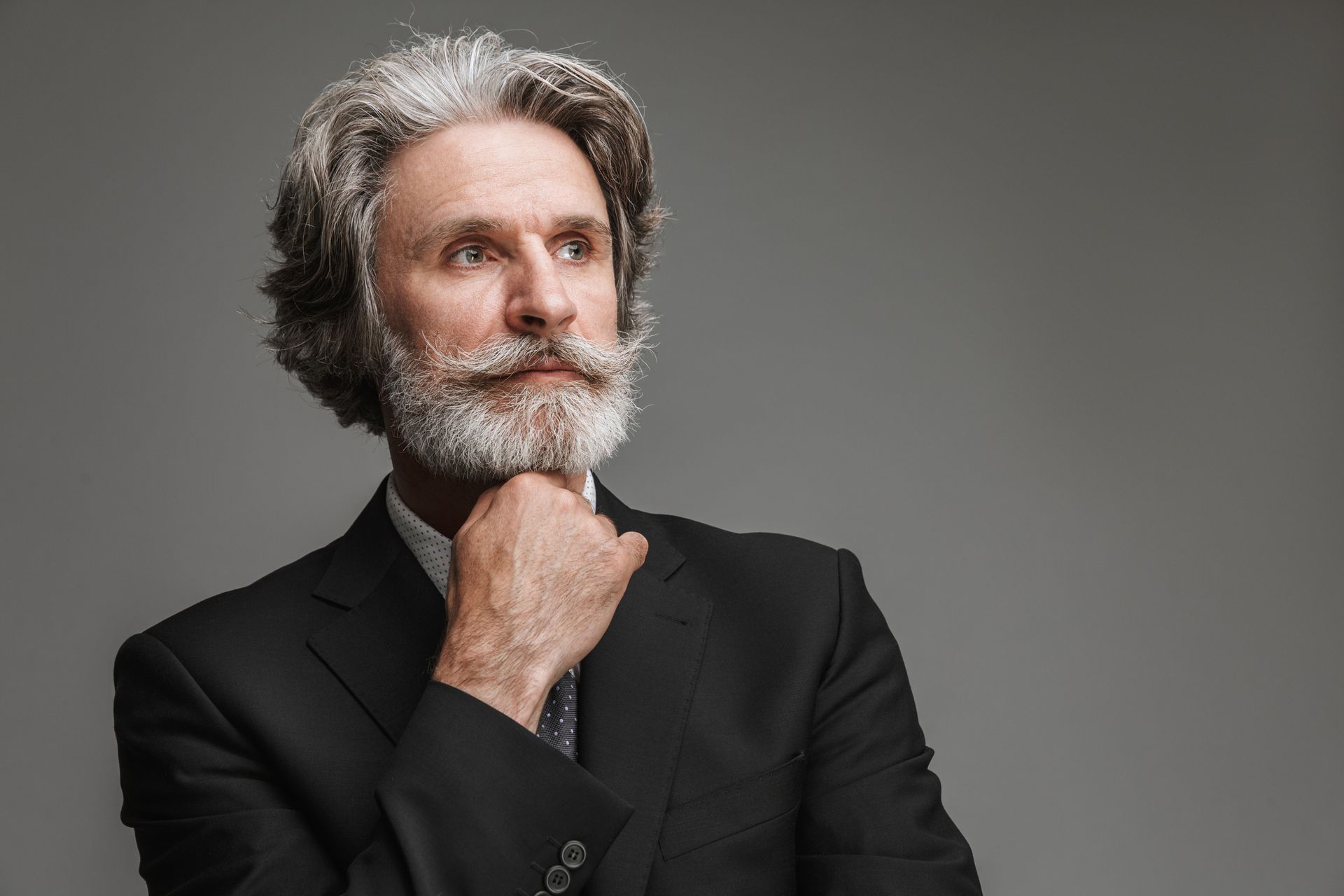 A person with a grey beard and wavy hair wears a suit and rests their chin on their hand, looking toward the right.