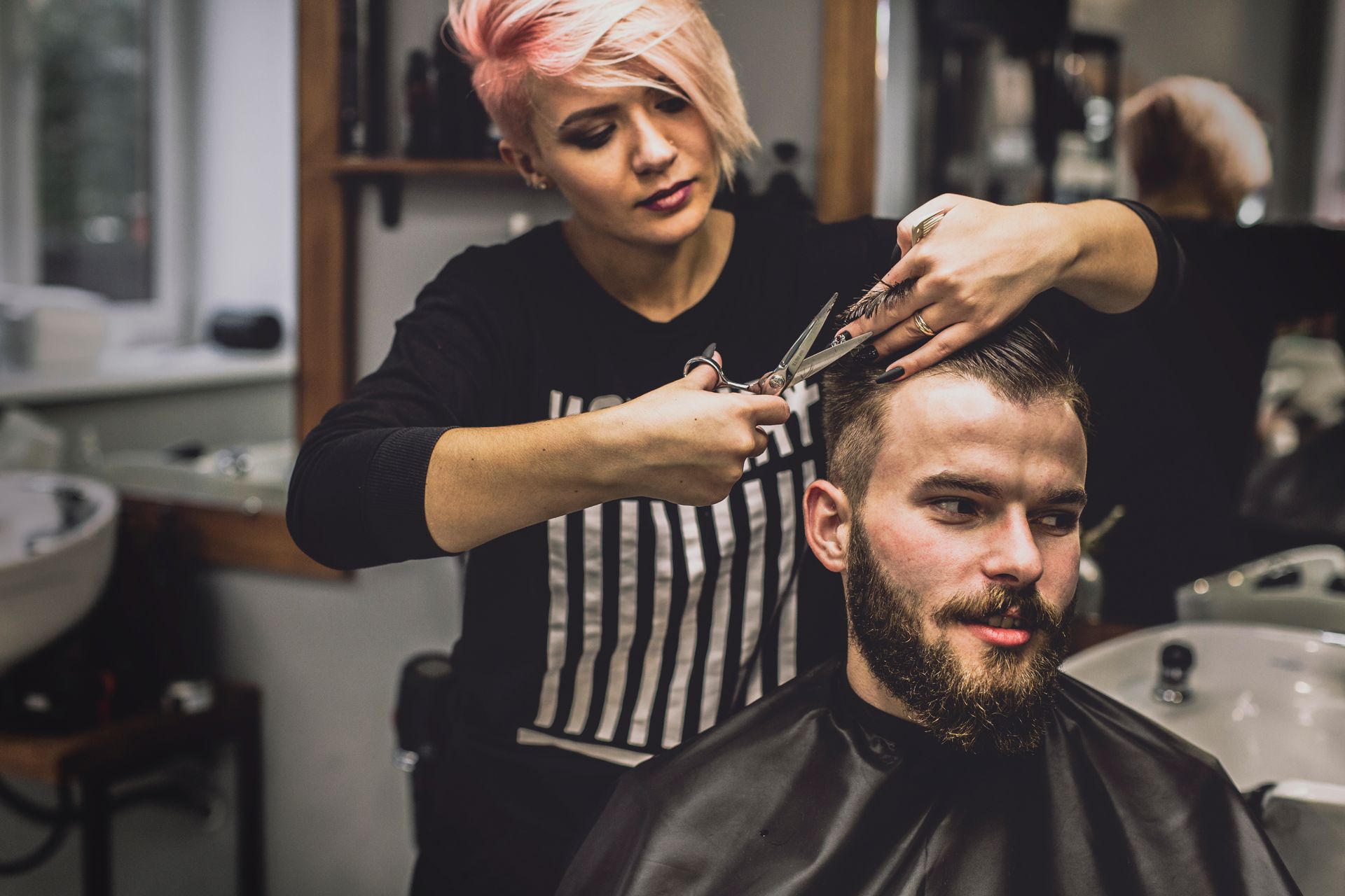 Hairdresser cutting a man's hair in a salon; she has pink hair, he wears a black cape.