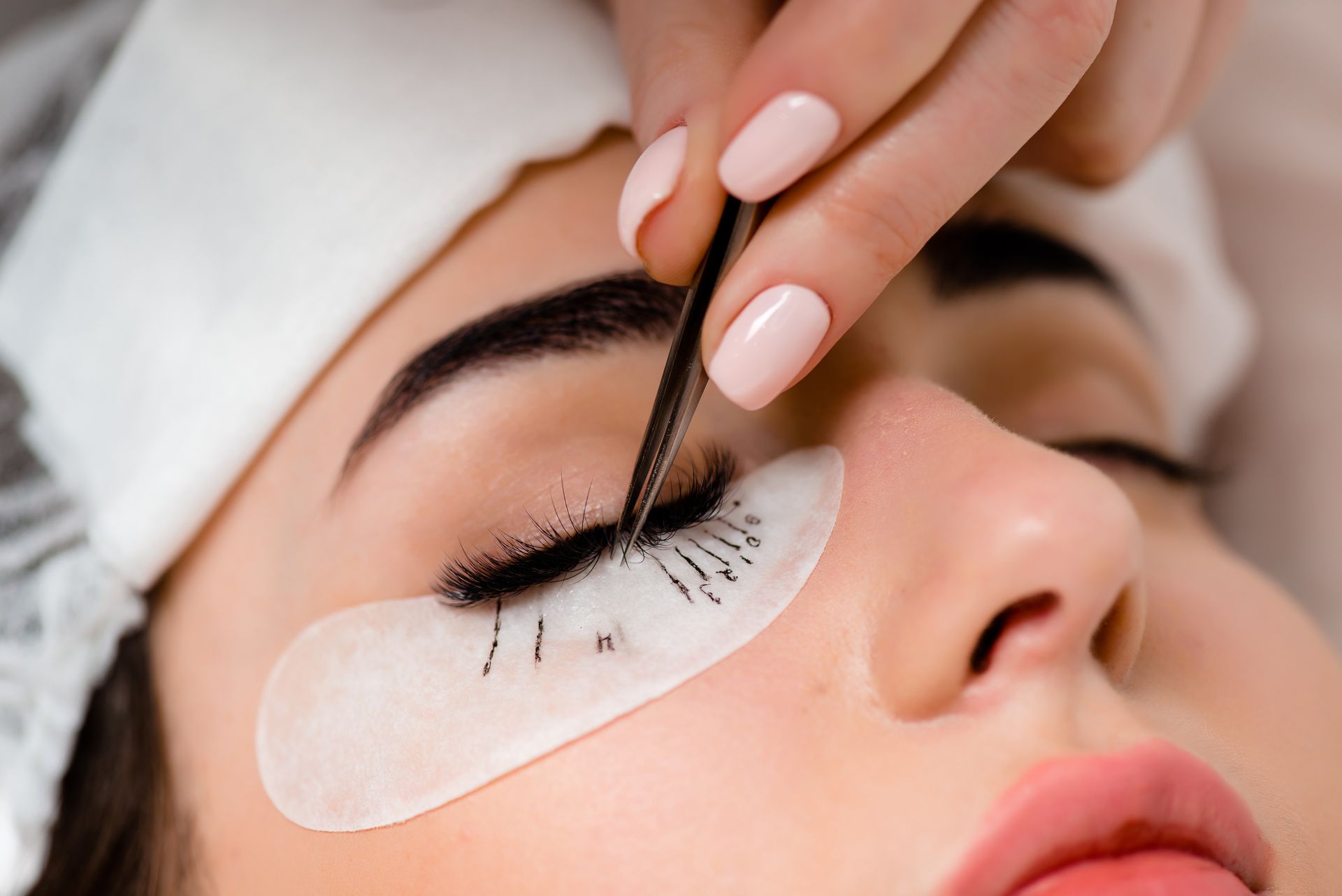 A person receiving eyelash extensions. A hand uses tweezers to apply a lash; eye closed, skin patches.