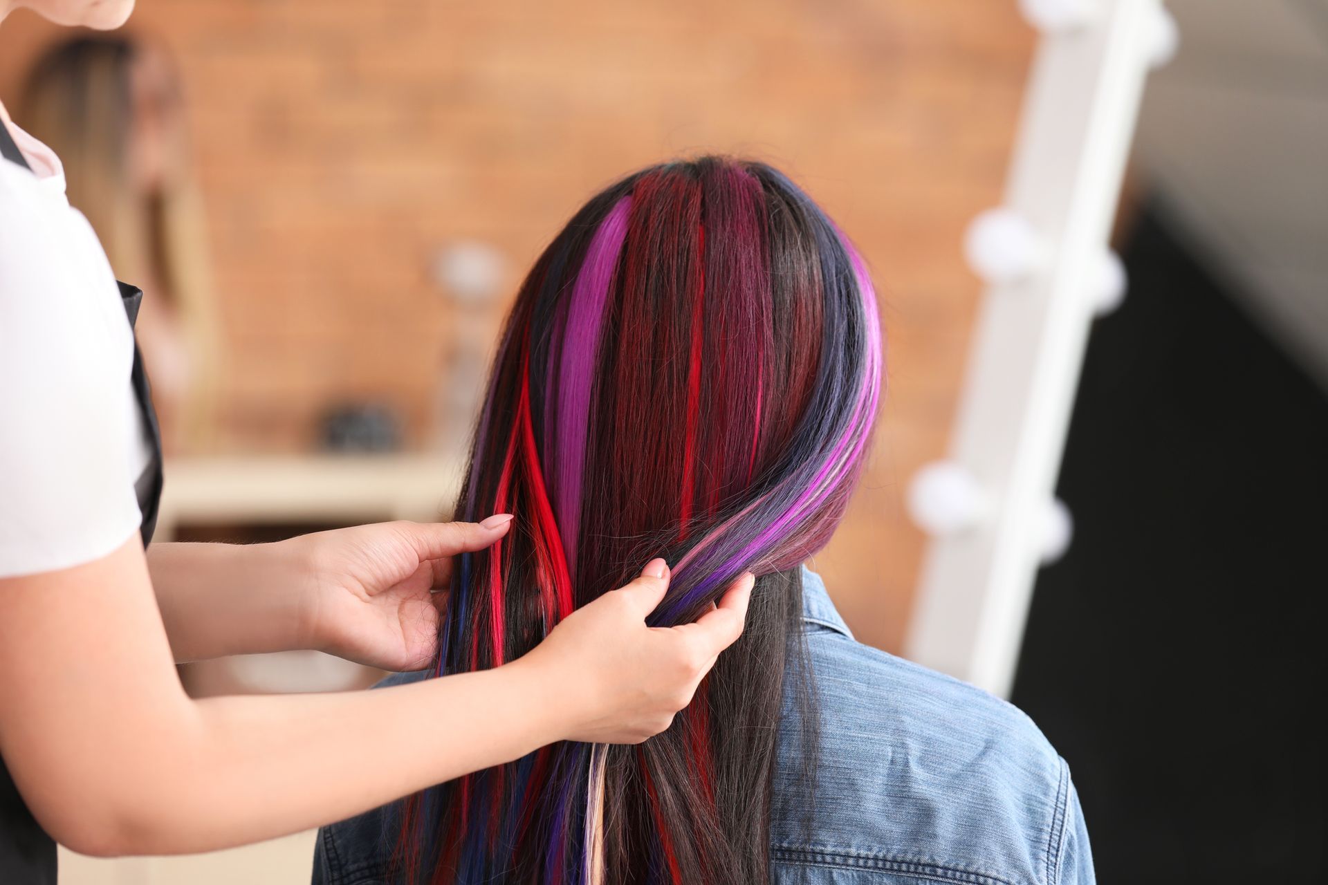 Person with dark hair and colorful streaks has hair styled by another.