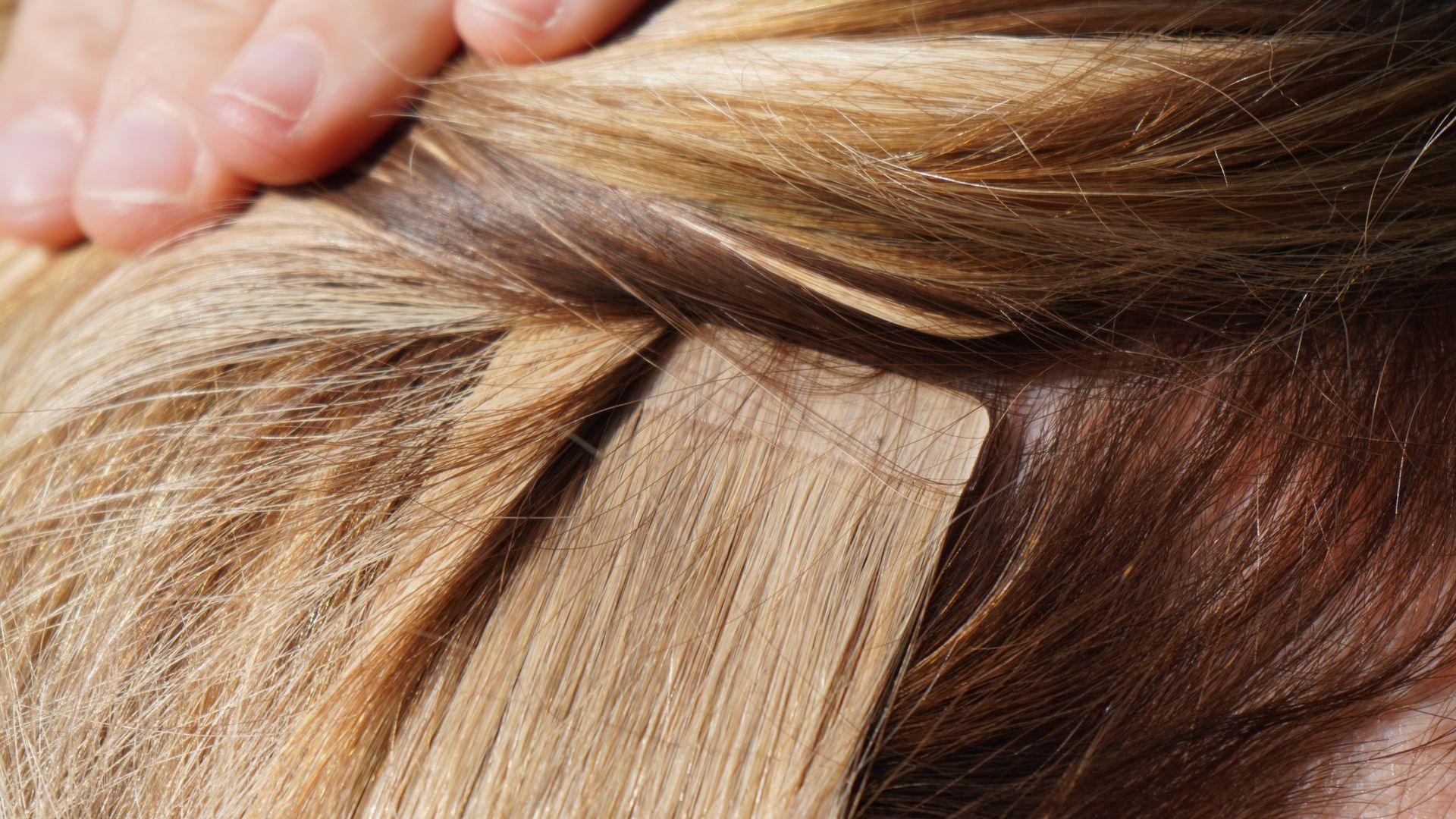 Blonde hair with tape-in extensions visible at the root. A hand touches the hair.