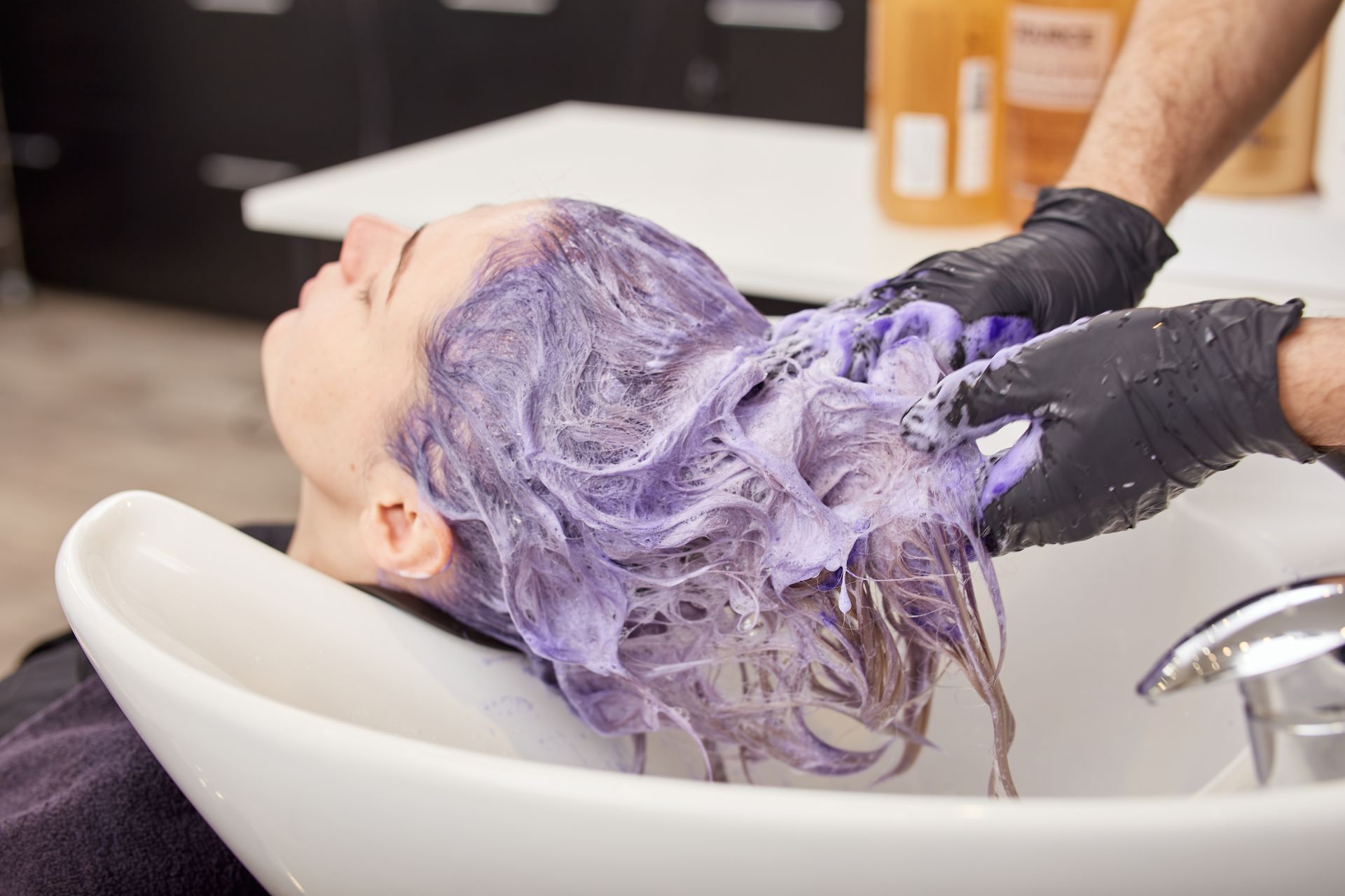 Person with purple-dyed hair has it washed in a salon sink by someone wearing black gloves.