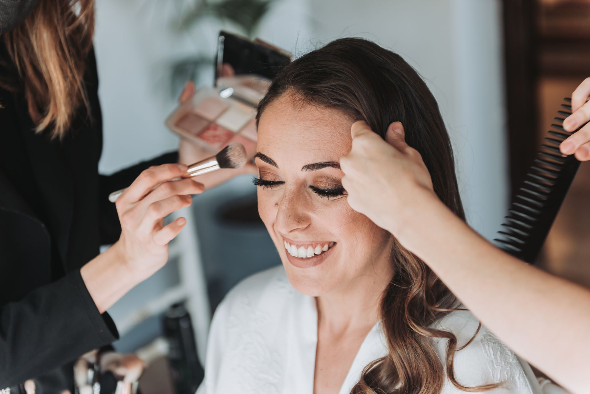 Woman having makeup and hair styled for an event. Smiling, with makeup artist holding brush, another holding comb.