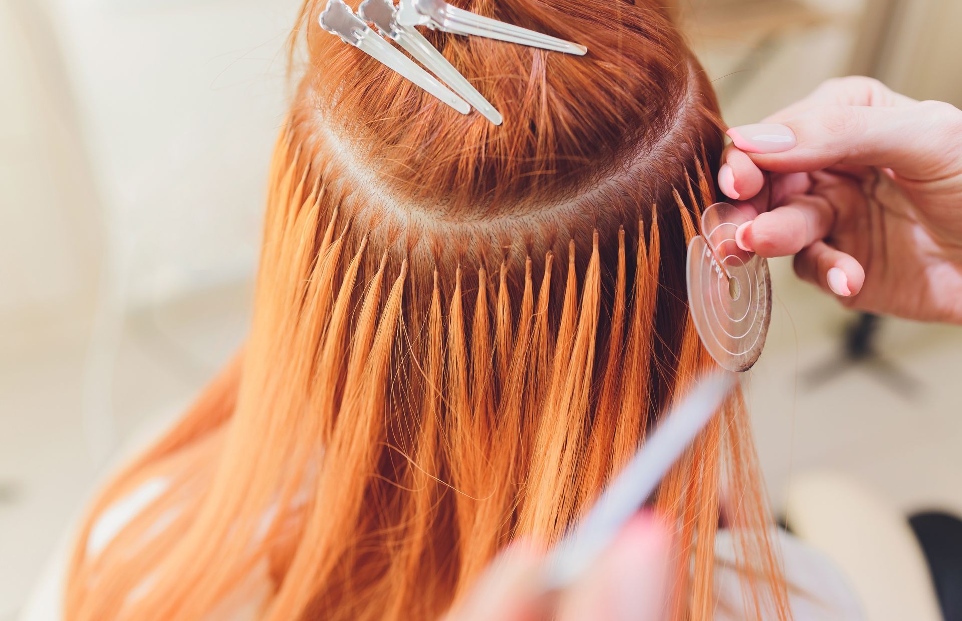 Hands attaching individual hair extensions to red hair near the scalp, with clips holding up other sections.