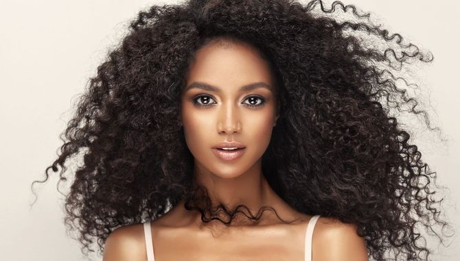 Woman with voluminous dark curly hair, pale skin, wearing a white top, looking directly at the viewer.