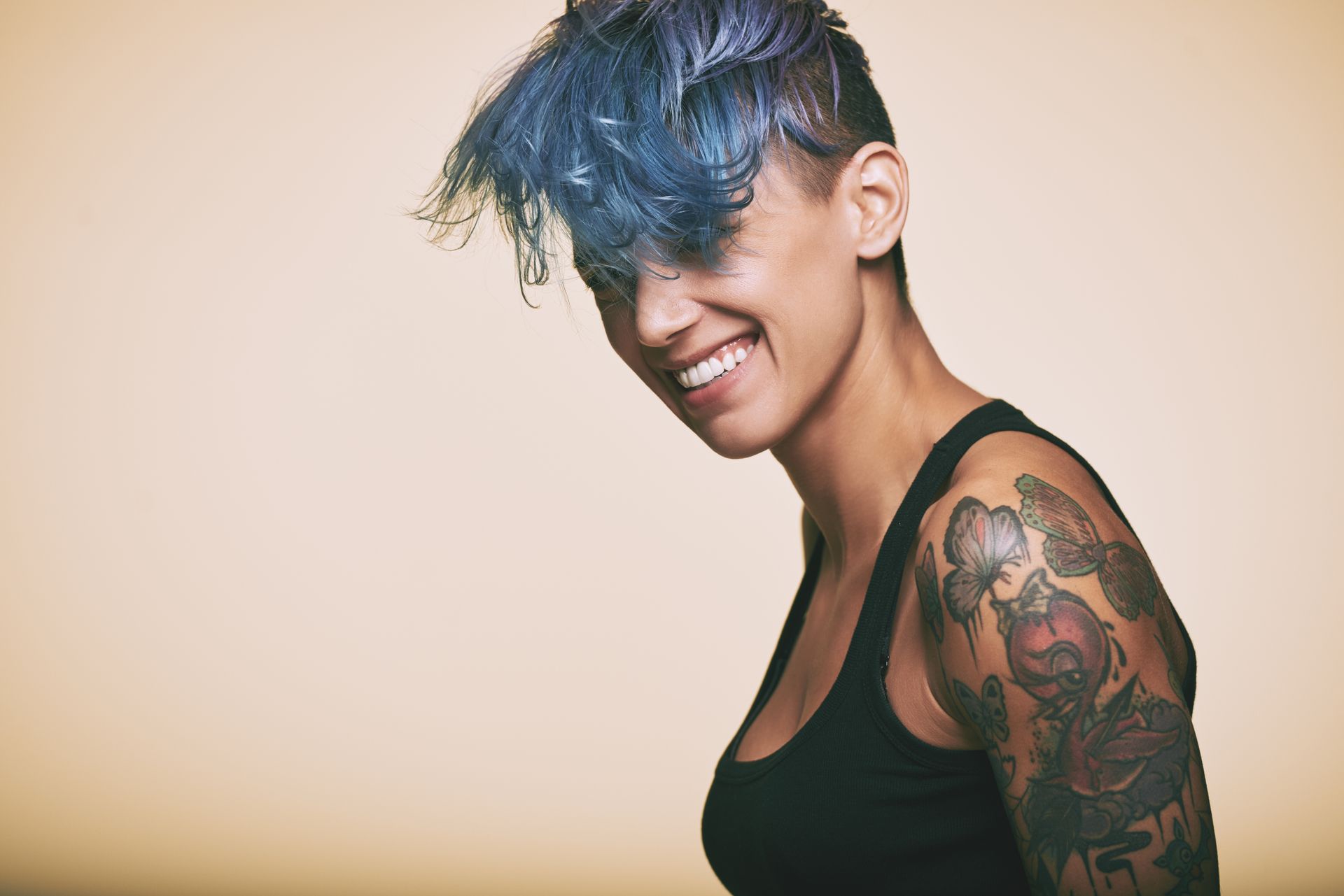 Woman with blue-dyed hair and arm tattoo smiles, wearing a black tank top, set against a beige backdrop.
