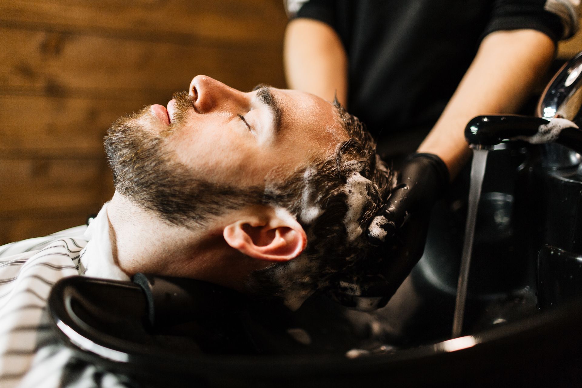 Person getting hair washed at salon. Water runs into sink as hands in black gloves lather hair with shampoo.
