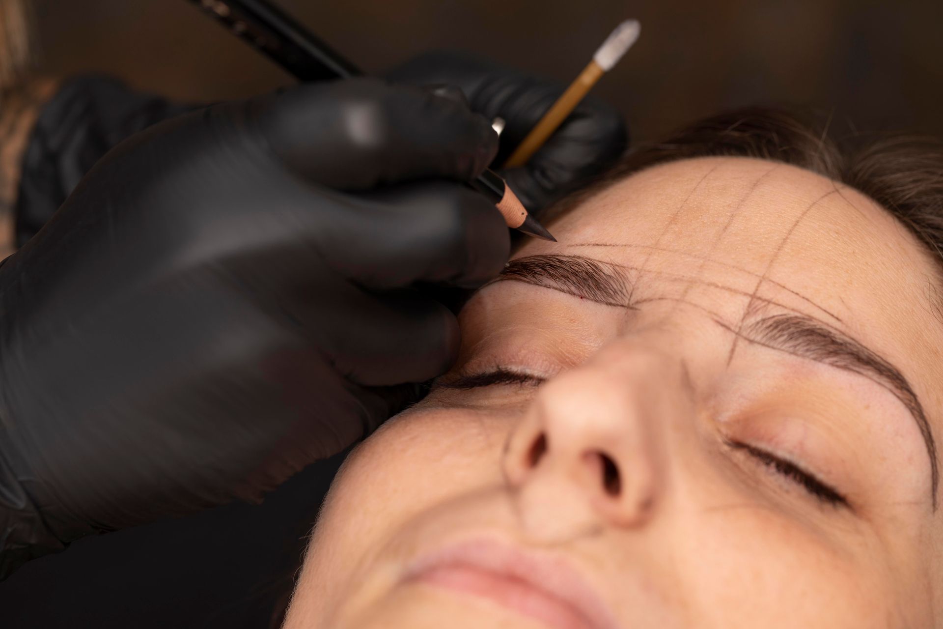 A person's eyebrows being shaped with a pencil by a person wearing black gloves; grid lines drawn.