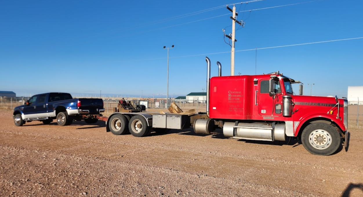 A red semi truck is towing a black truck in a dirt field.