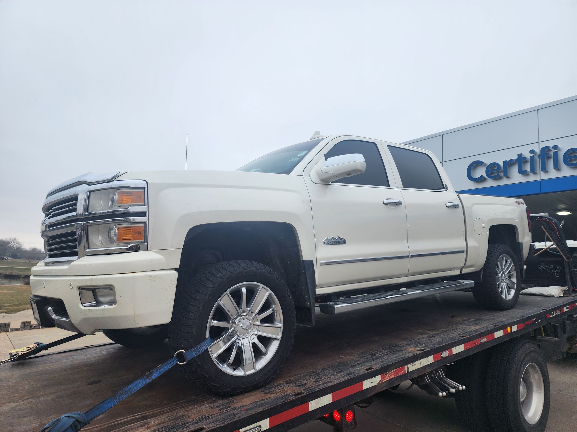 A white pickup truck secured with blue tie-down straps on the bed of a flatbed tow truck.