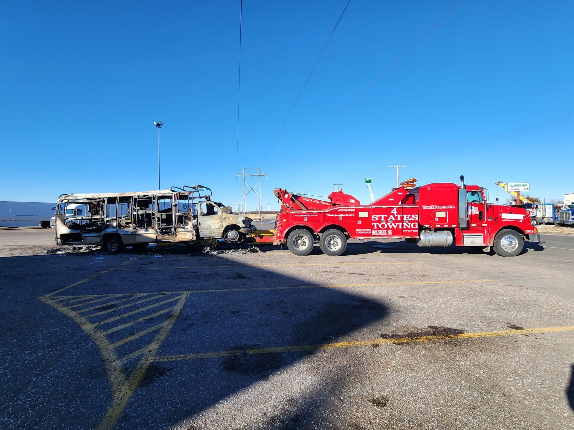 A bright red tow truck hauls a severely fire-damaged, charred vehicle across an empty, sunlit paved lot.