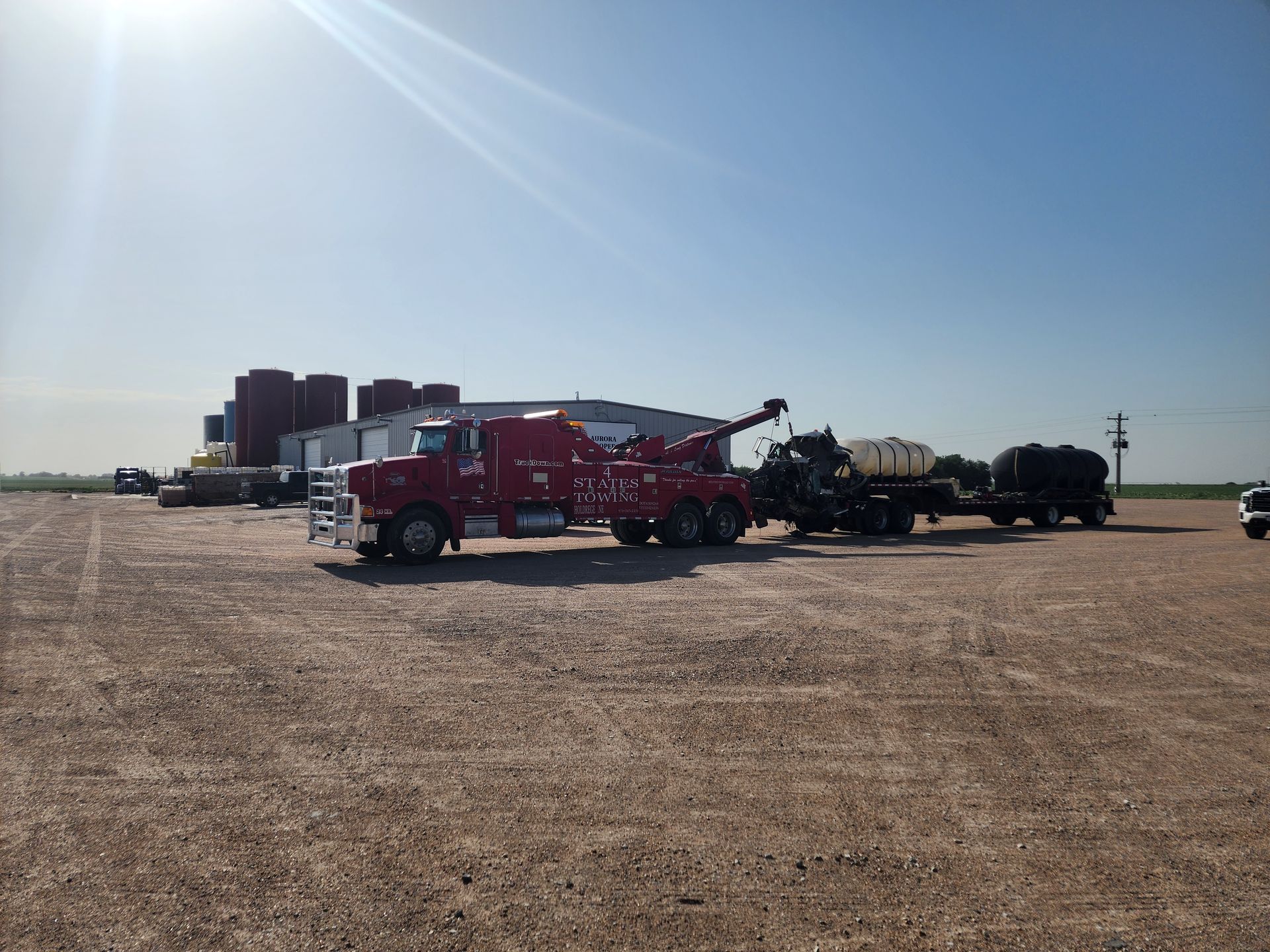 A red heavy-duty tow truck is pulling a trailer with white agricultural tanks across a large, sunlit gravel lot.