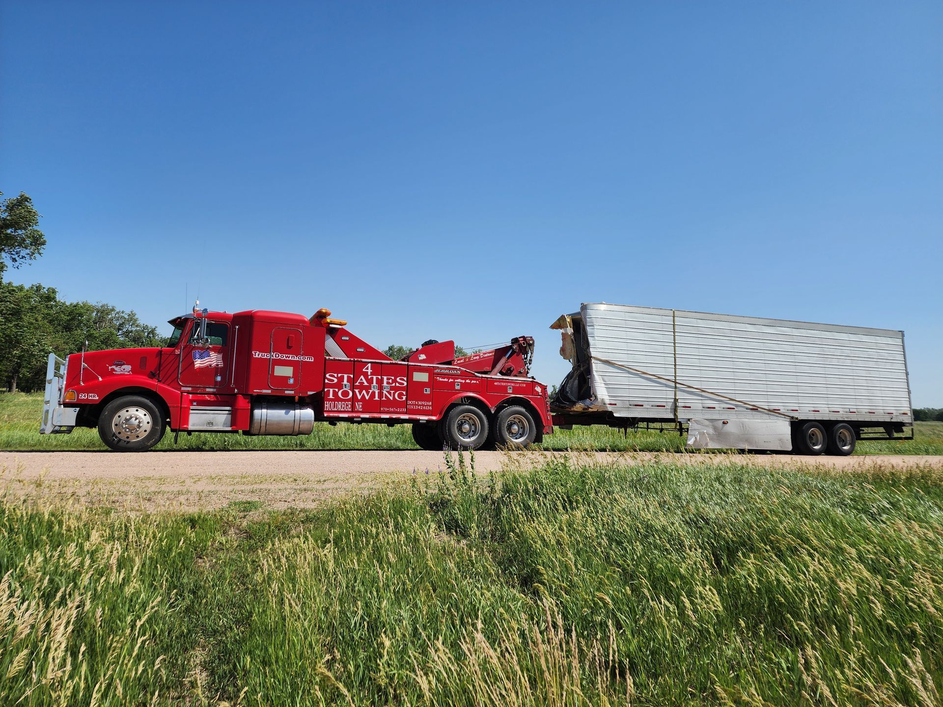 A bright red tow truck pulling a silver cargo trailer along a rural road against a clear blue sky.