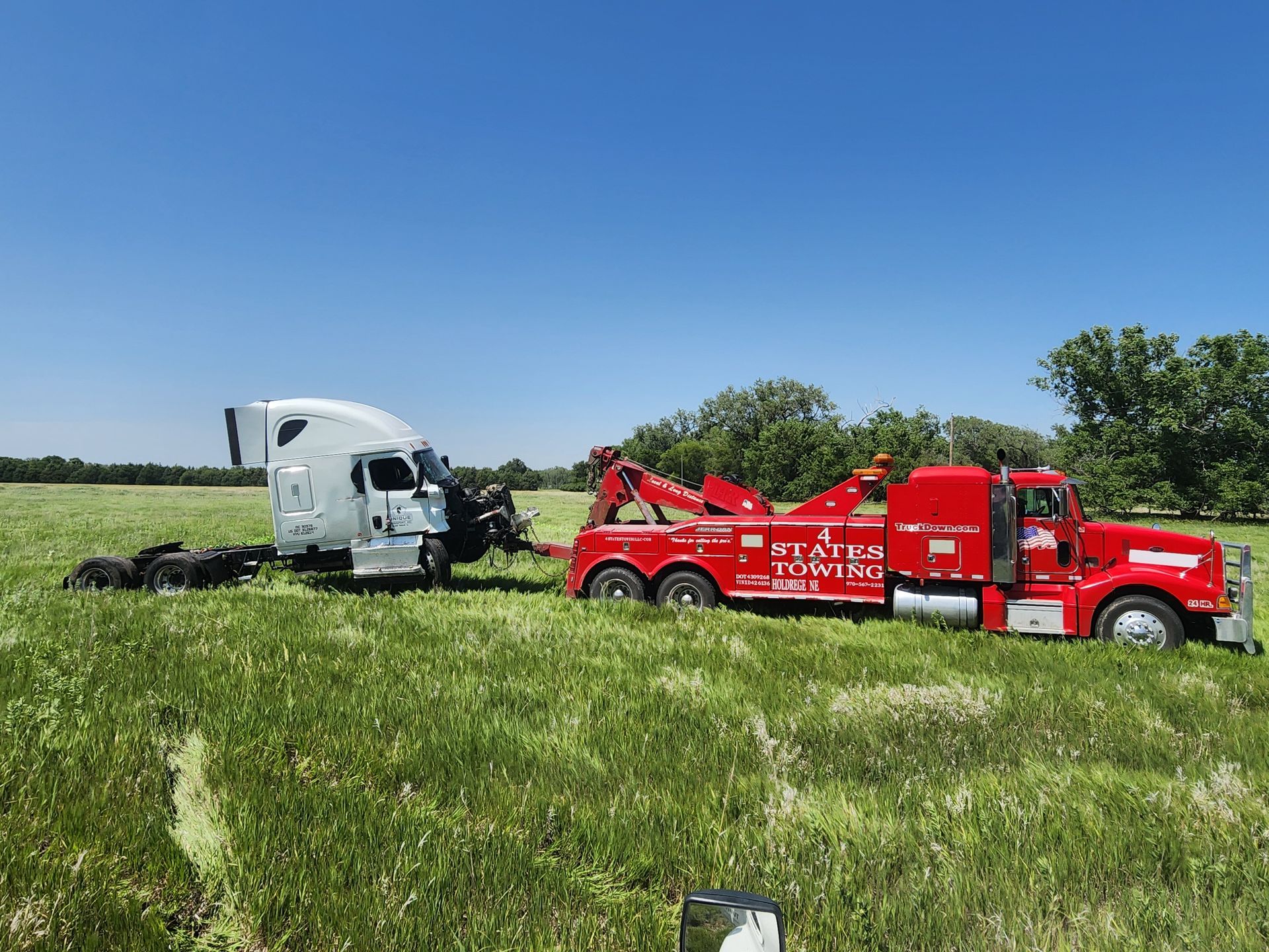 A bright red tow truck pulls a damaged white semi-truck cab through a grassy, sunlit field.