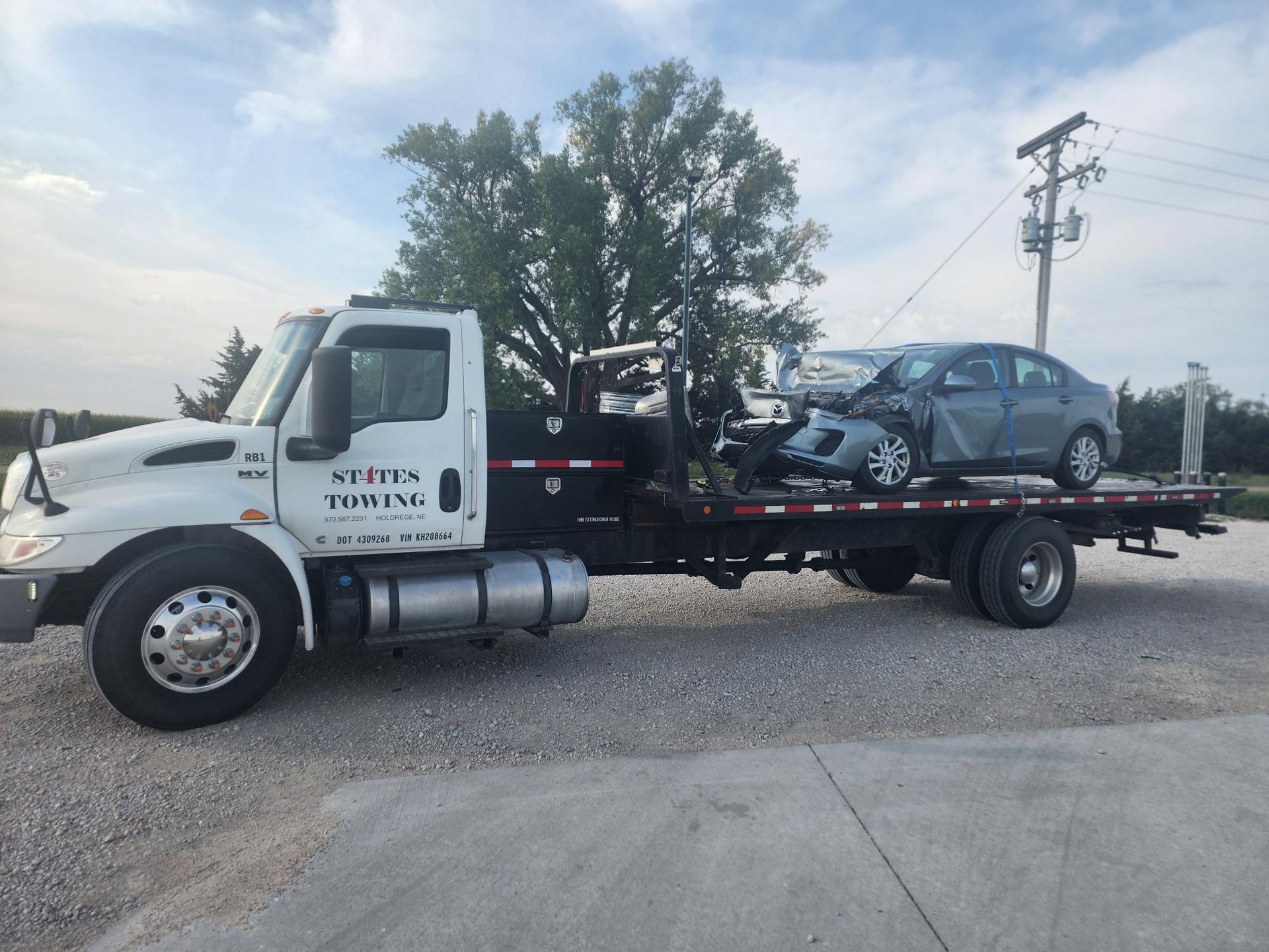 A white Stites Towing flatbed truck hauling a heavily damaged grey sedan on a gravel lot under a blue sky.