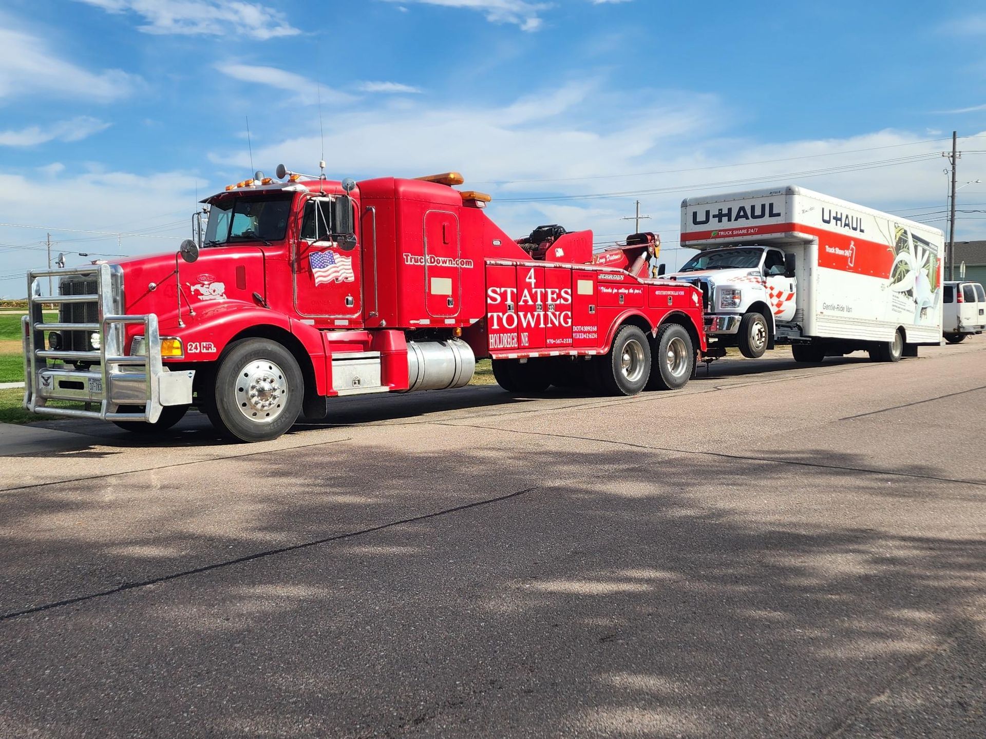 A red Status Towing heavy-duty tow truck pulling a white U-Haul moving truck along a paved road under a blue sky.