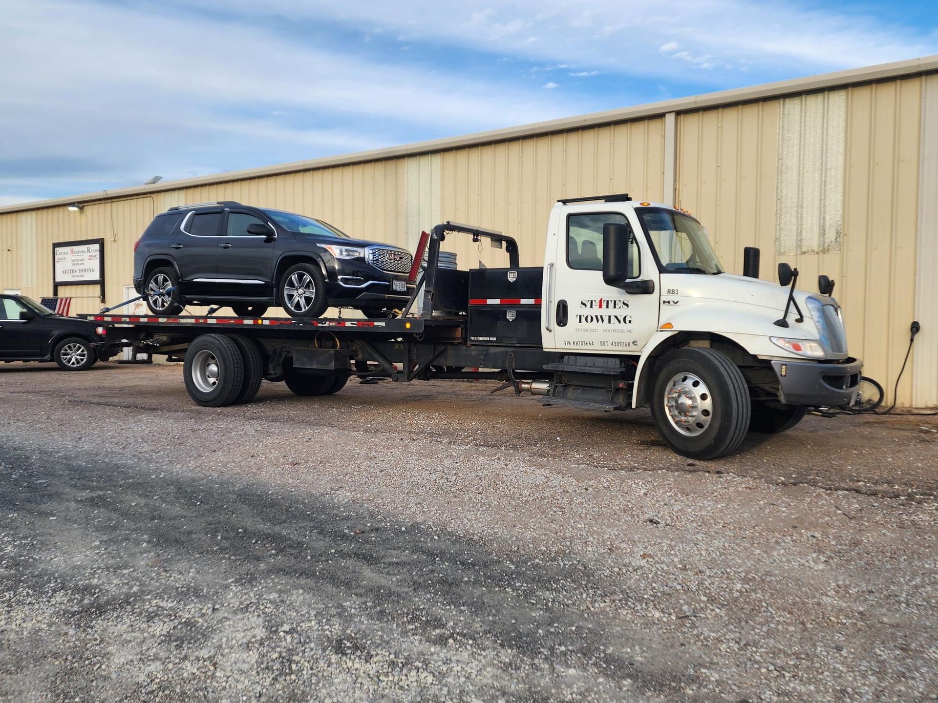A white flatbed tow truck carrying a dark SUV parked on a gravel lot in front of a tan metal building.