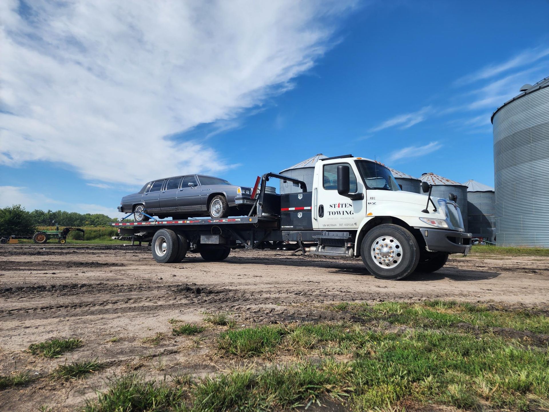 A grey limousine loaded onto a white flatbed tow truck on a dirt lot with large silver grain silos under a blue sky.