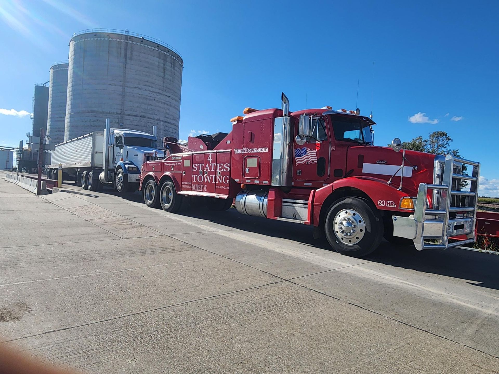 A red heavy-duty tow truck is hooked up to a white semi-truck and trailer near large industrial silos on a sunny day.