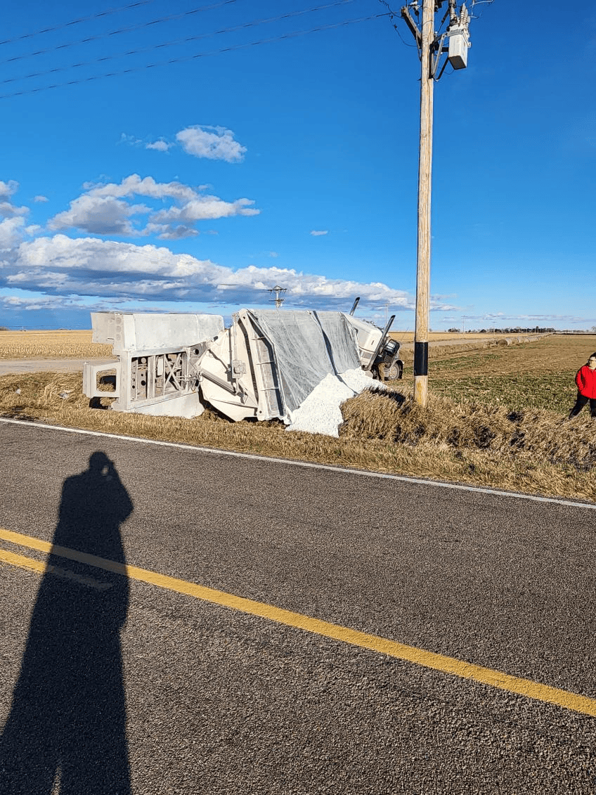 A wrecked vehicle lies in a rural field next to a utility pole beside an asphalt road under a bright blue sky.