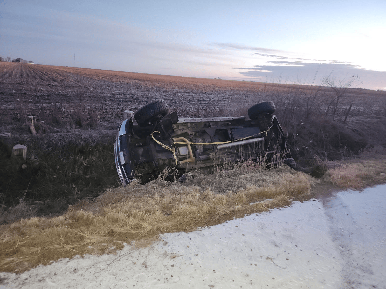A dark-colored pickup truck overturned in a grassy ditch next to a dirt road and a harvested field.