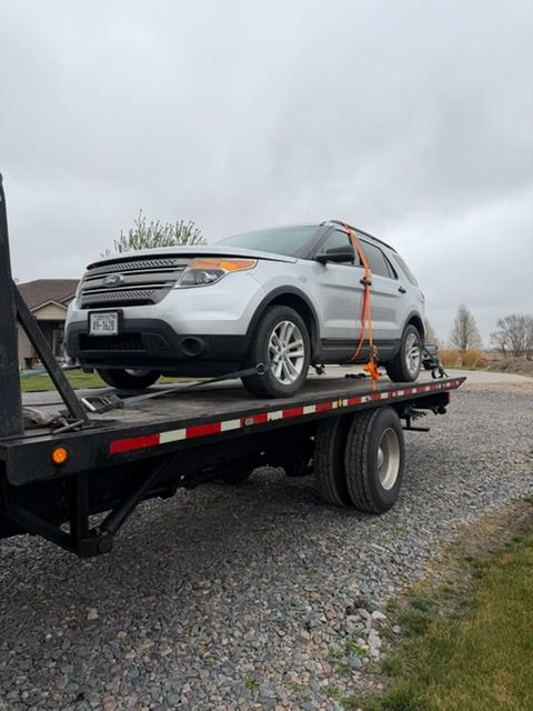 Silver SUV strapped to a flatbed trailer on a gravel road under a cloudy sky