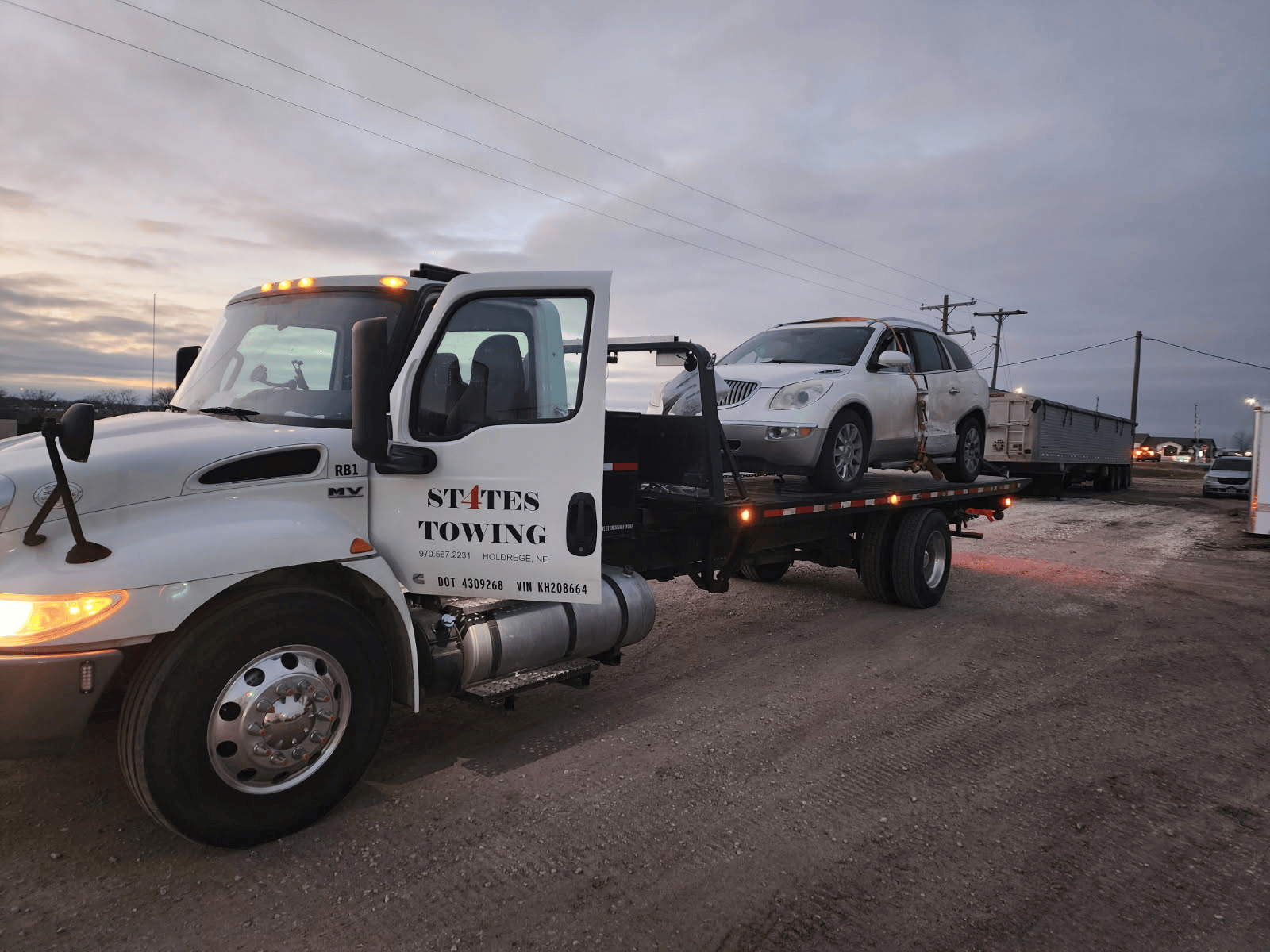 A white States Towing flatbed truck hauling a white SUV parked on a dirt lot under a cloudy sky.