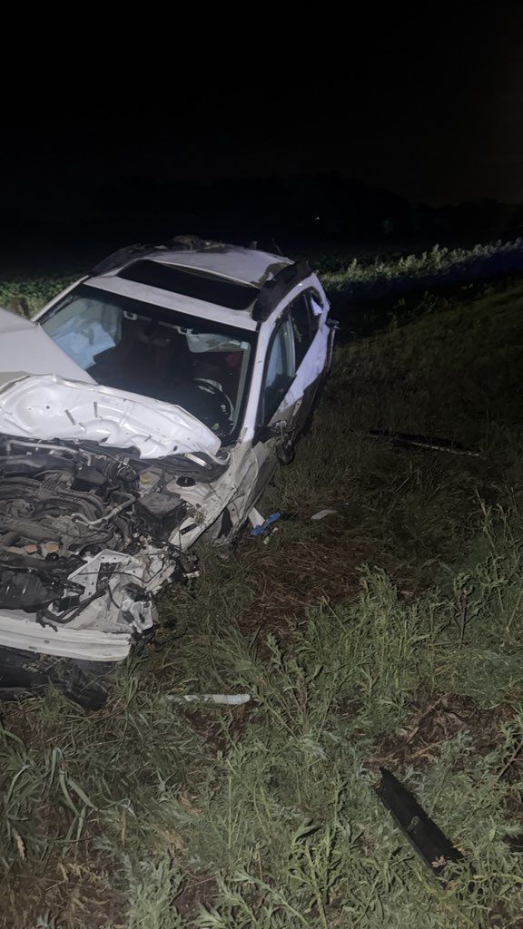 Nighttime crash scene with a damaged white car off the road in a grassy ditch.