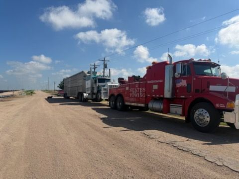 Red tow truck and semi-trucks parked on a dirt roadside under a blue sky with clouds