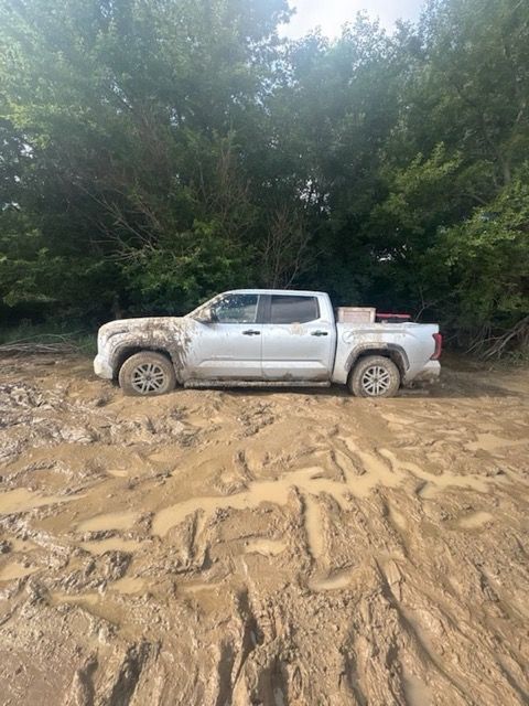 White pickup truck stuck in muddy terrain with trees in the background