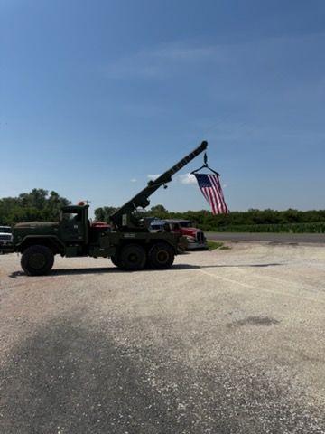 Military tow truck raising a large U.S. flag in an open paved lot under a clear blue sky