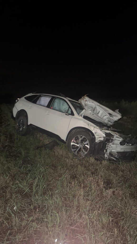 White SUV wrecked off-road at night, front end heavily damaged in a grassy field.