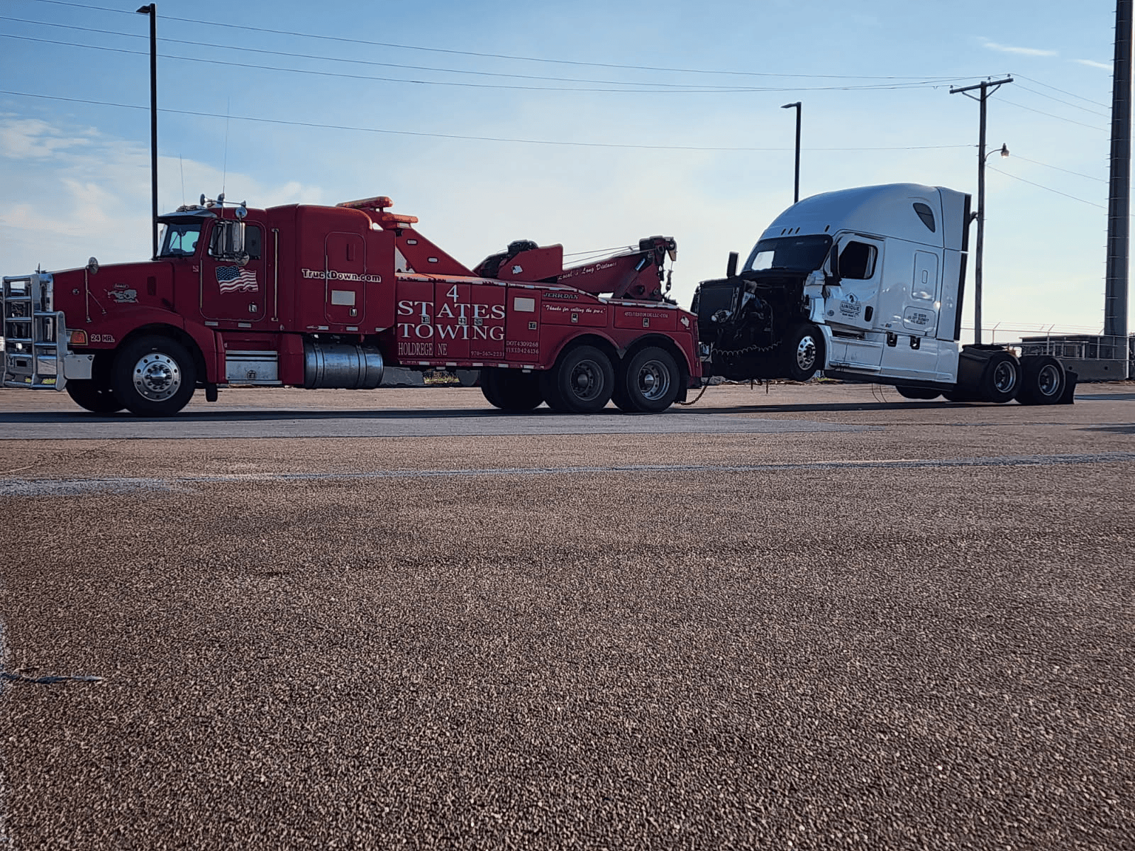 A red heavy-duty tow truck is towing a damaged white semi-truck in a large, gravel-covered parking lot.