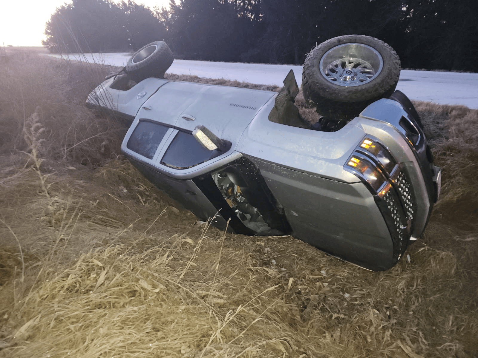 A silver pickup truck lying on its side in a grassy ditch next to a road.