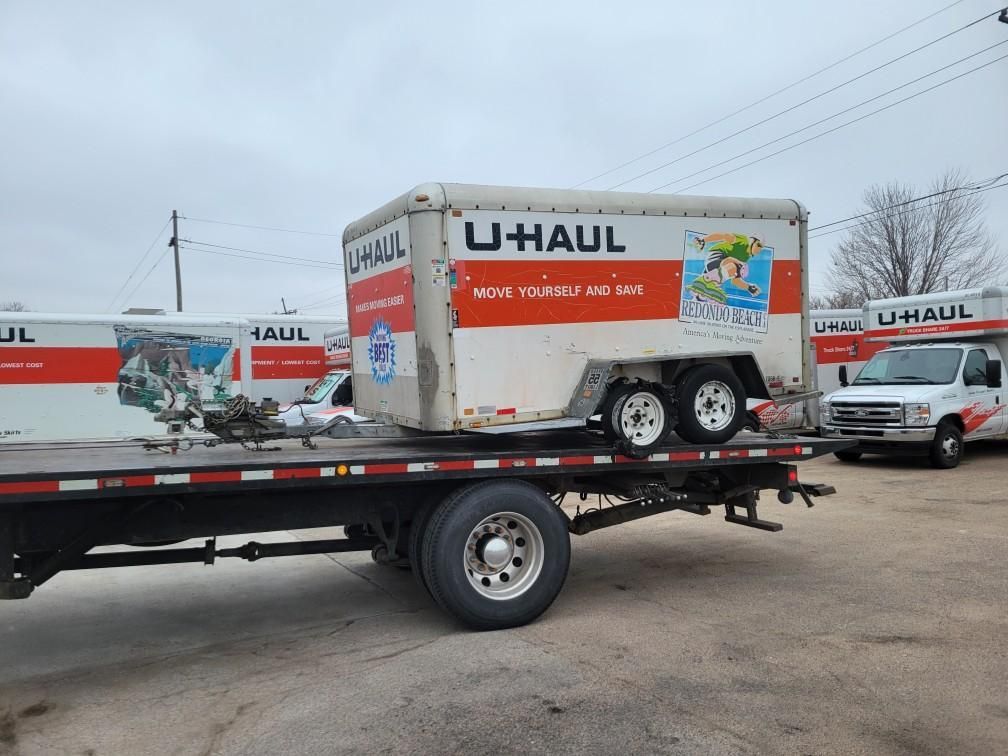 U-Haul trailer loaded on a flatbed truck in a snowy lot, with more U-Haul trucks parked nearby.