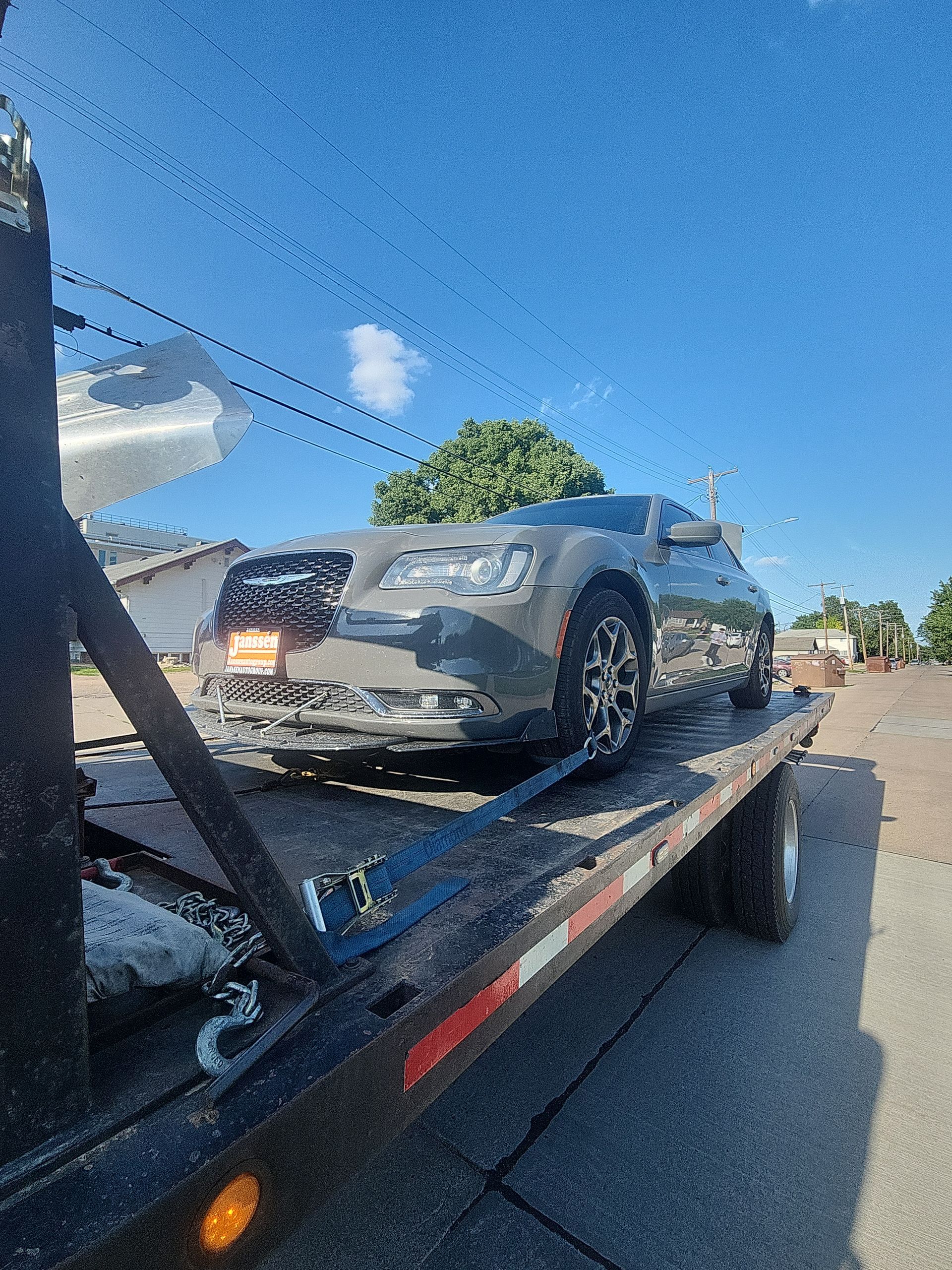 A gray Chrysler 300 sedan sits secured with blue tie-down straps on a flatbed tow truck parked on a sunny street.