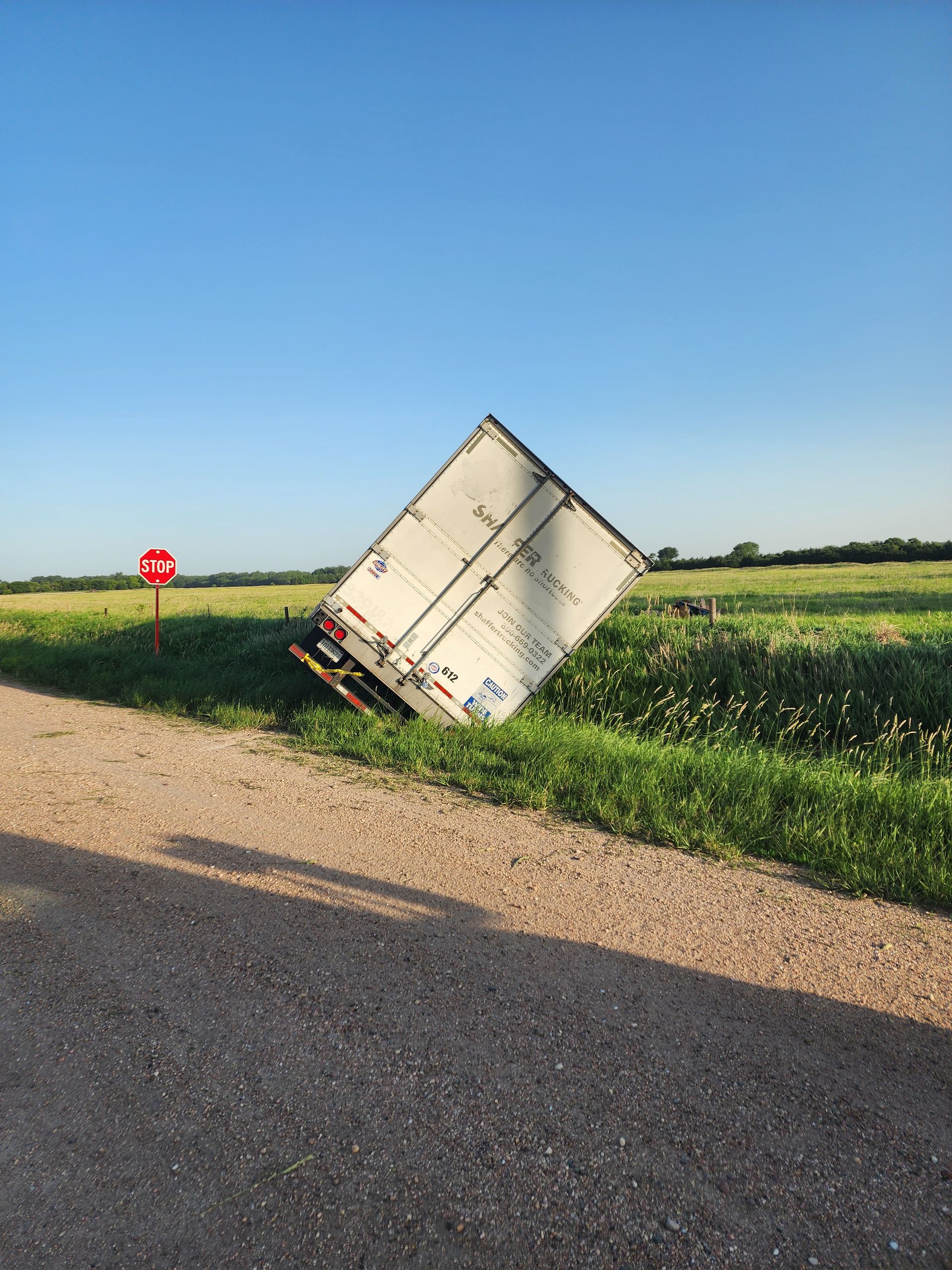 A white shipping container sits at a tilted angle in a grassy field next to a gravel road with a red stop sign nearby.