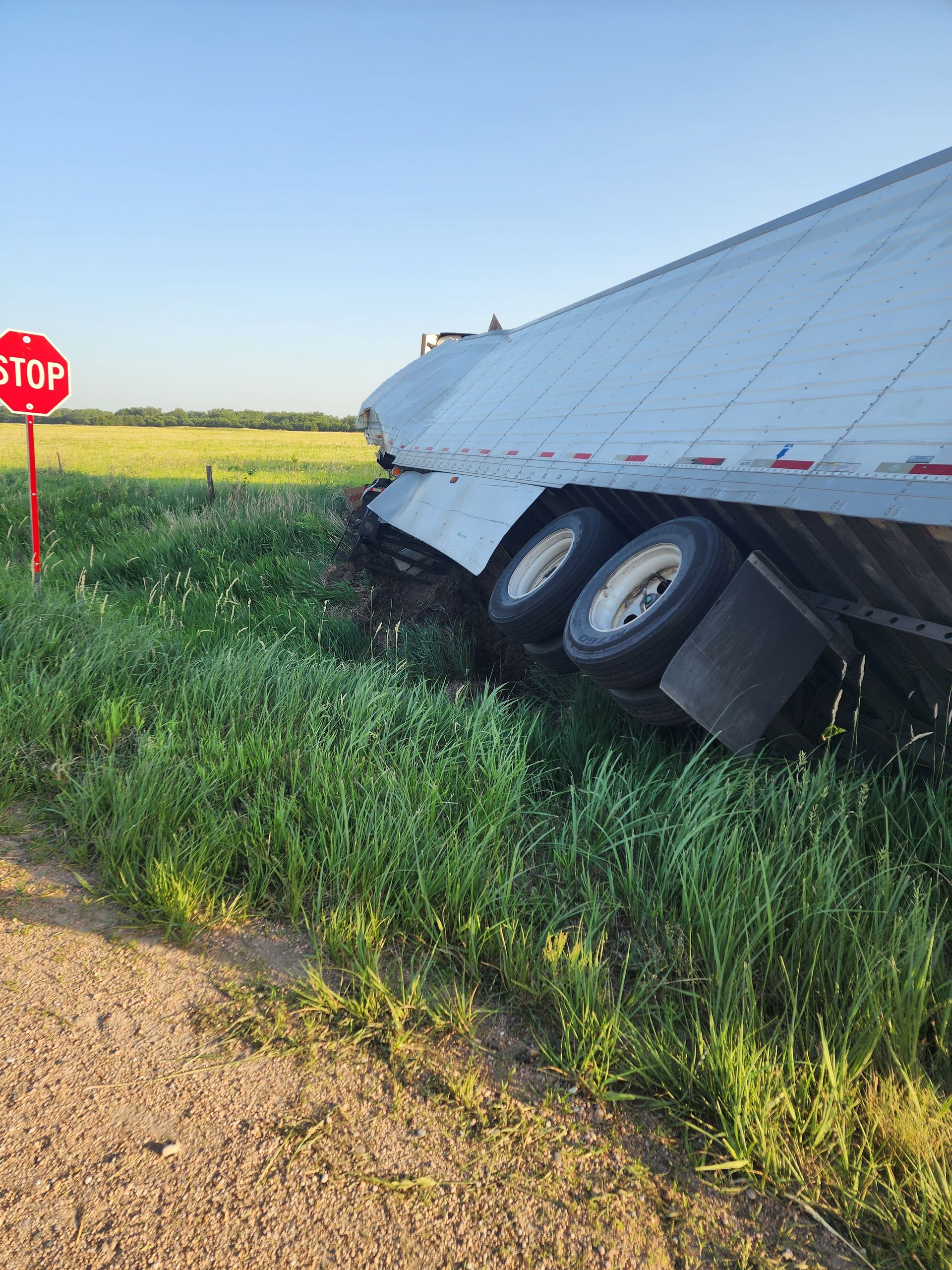 A large semi-trailer truck has veered off the road and is tilted into a grassy ditch near a red stop sign.