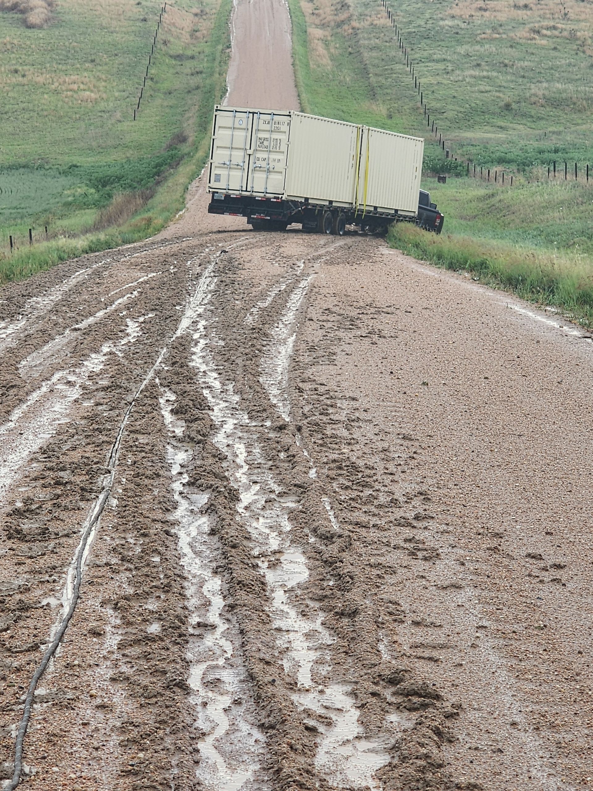 A semi-trailer truck is jackknifed across a muddy, rural dirt road flanked by grassy hills.