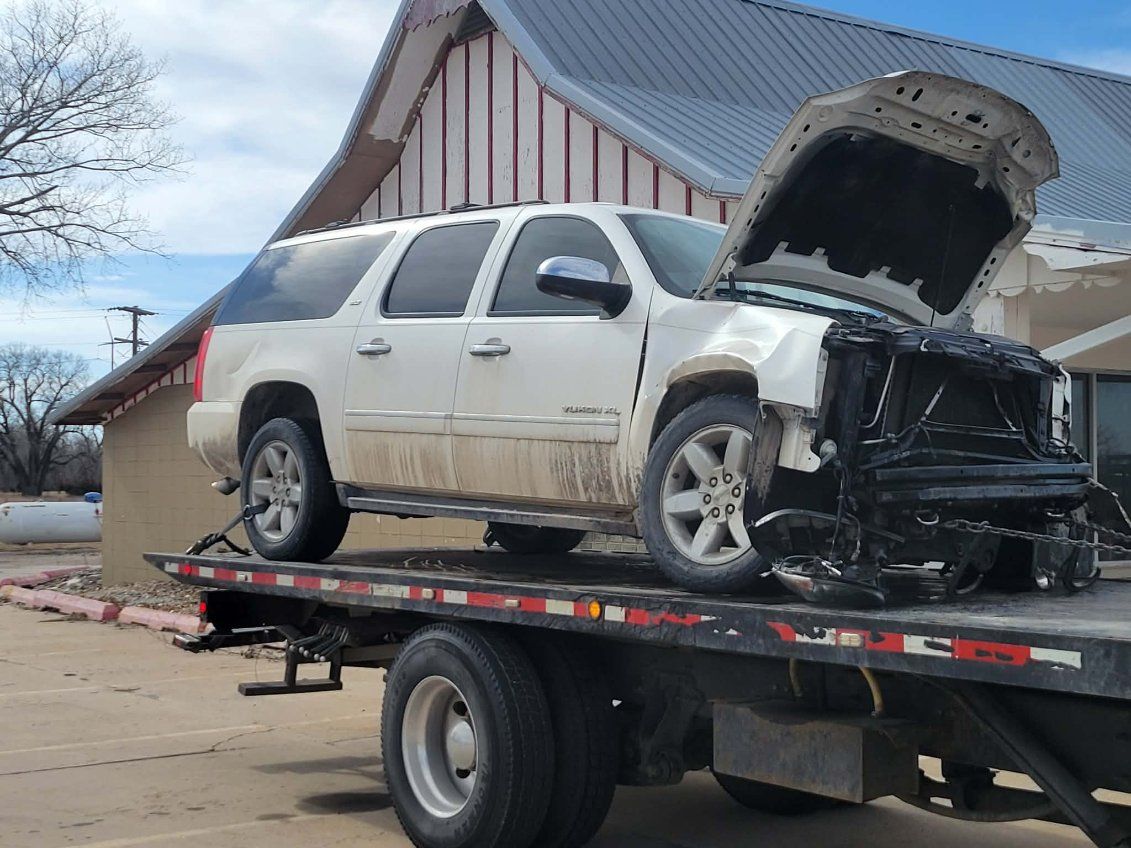 A white SUV with significant front-end damage and an open hood sits on a flatbed tow truck.