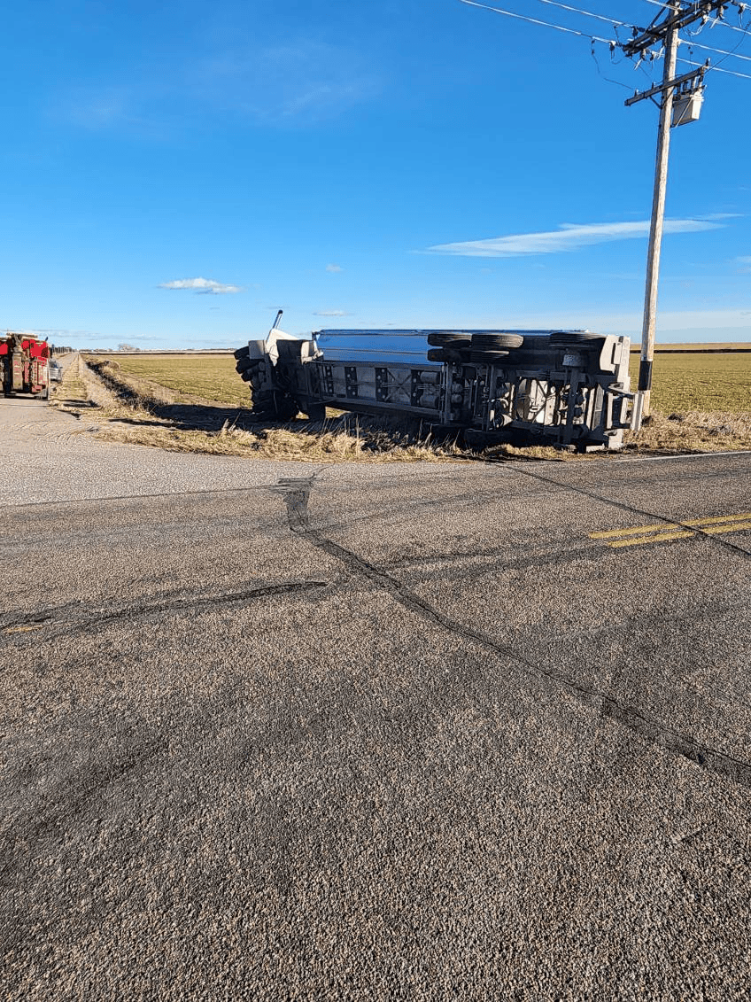 A white semi-truck overturned on its side in a field beside a paved rural road and a utility pole.