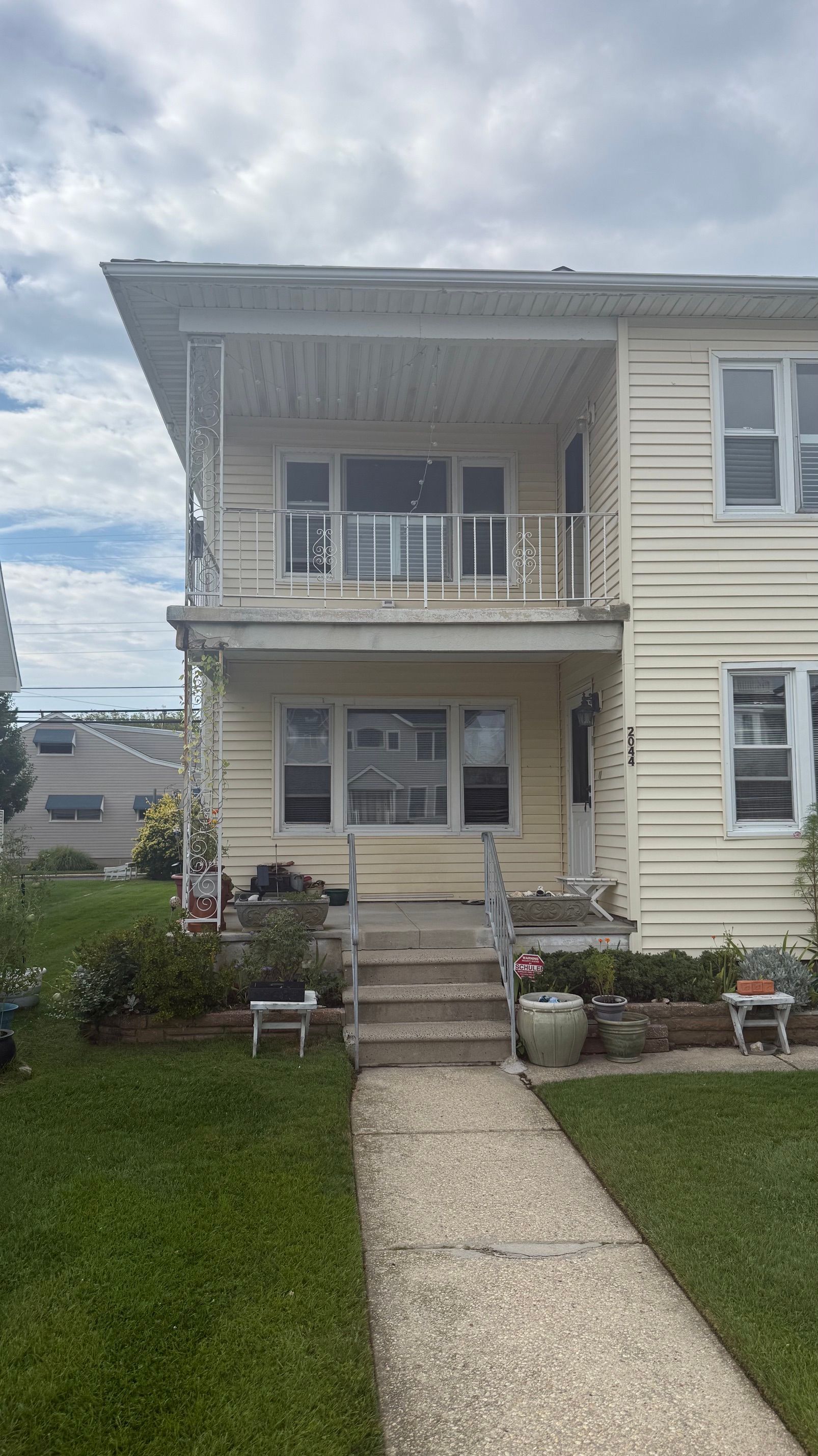 Two-story beige house with a small balcony, front steps, and a concrete walkway leading through a lawn.