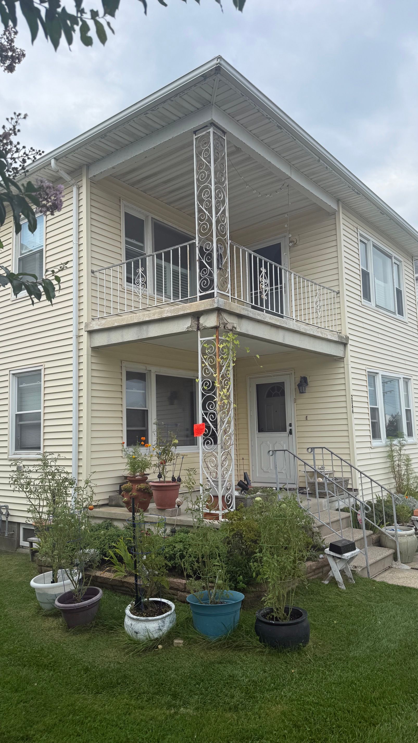 Two-story beige house with a front porch, white trim, and potted plants in the yard.