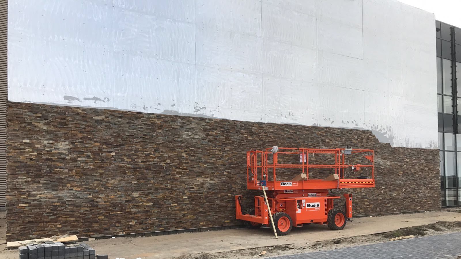 Een oranje schaarlift staat geparkeerd voor een gebouw in aanbouw.