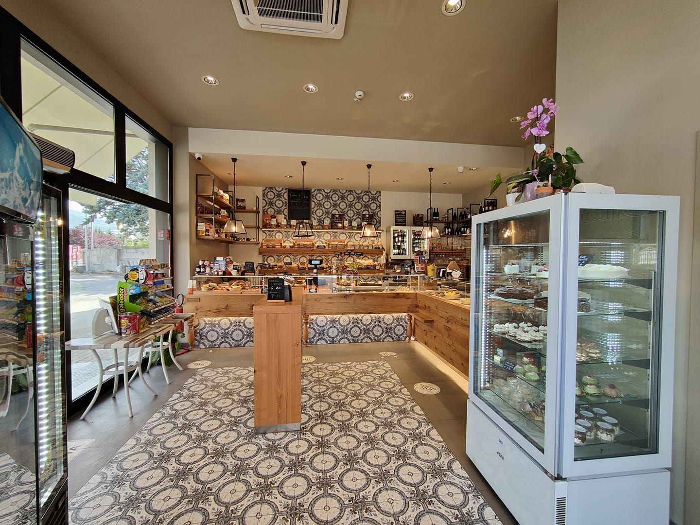 Interno di una panetteria con pavimento piastrellato decorato, bancone in legno, scaffali espositivi e vetrina refrigerata.
