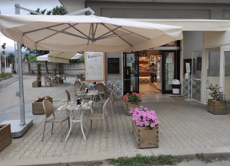 Dehor esterno del caffè con tavoli e sedie sotto un grande ombrellone, decorato con fiori rosa in vasi di legno.